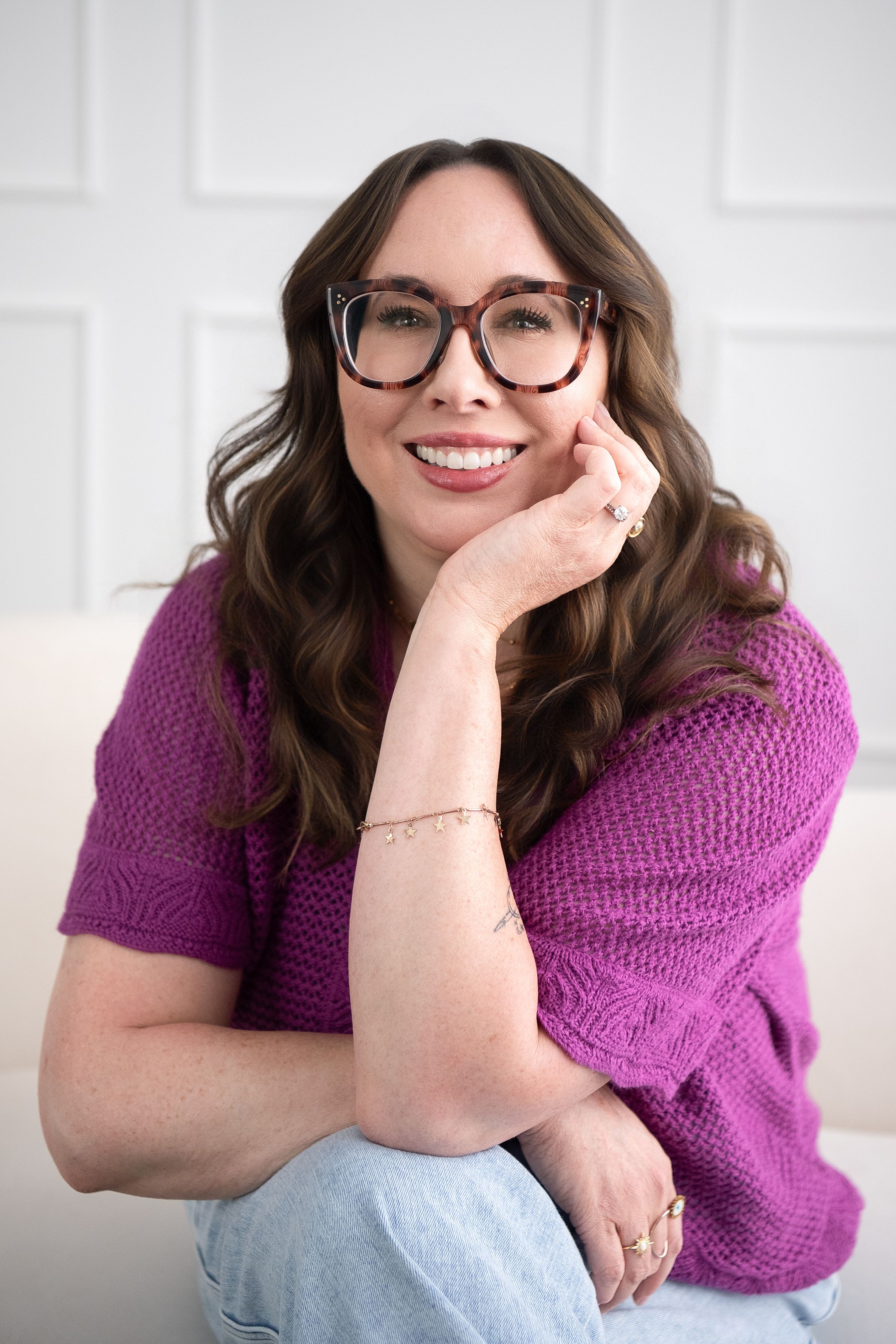Smiling woman with glasses and wavy brown hair wearing a purple knit sweater, posing casually on a white sofa.