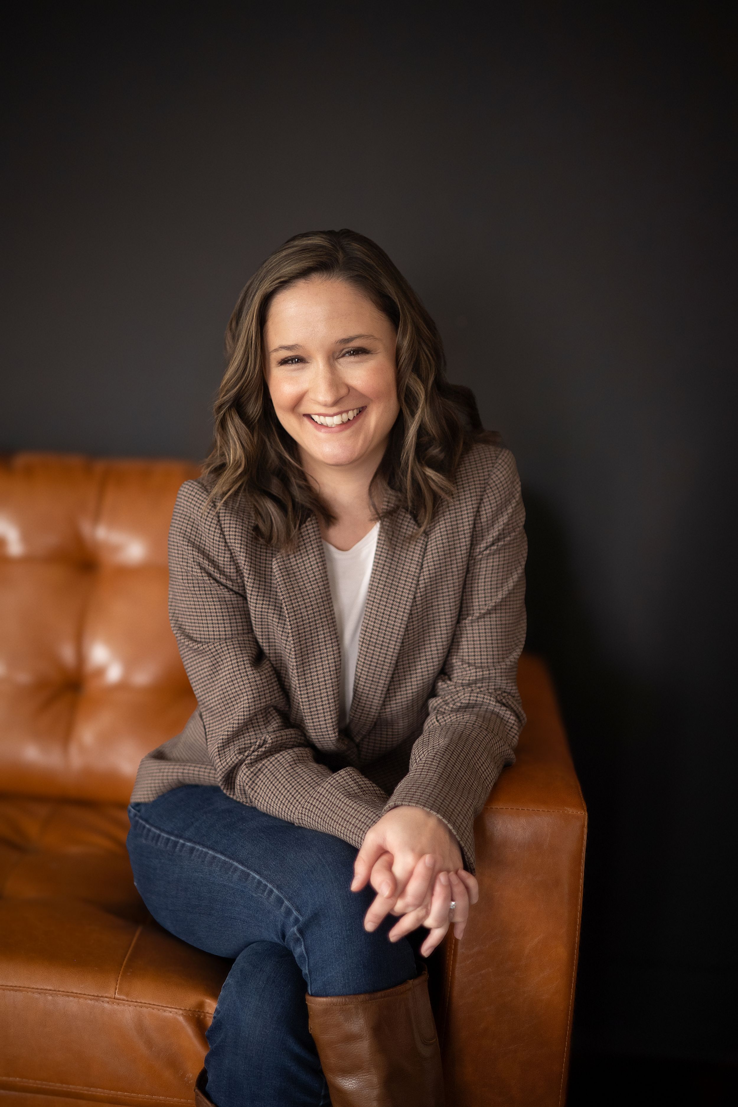 Smiling woman in brown blazer and jeans sitting on a tan leather couch against a dark gray wall.