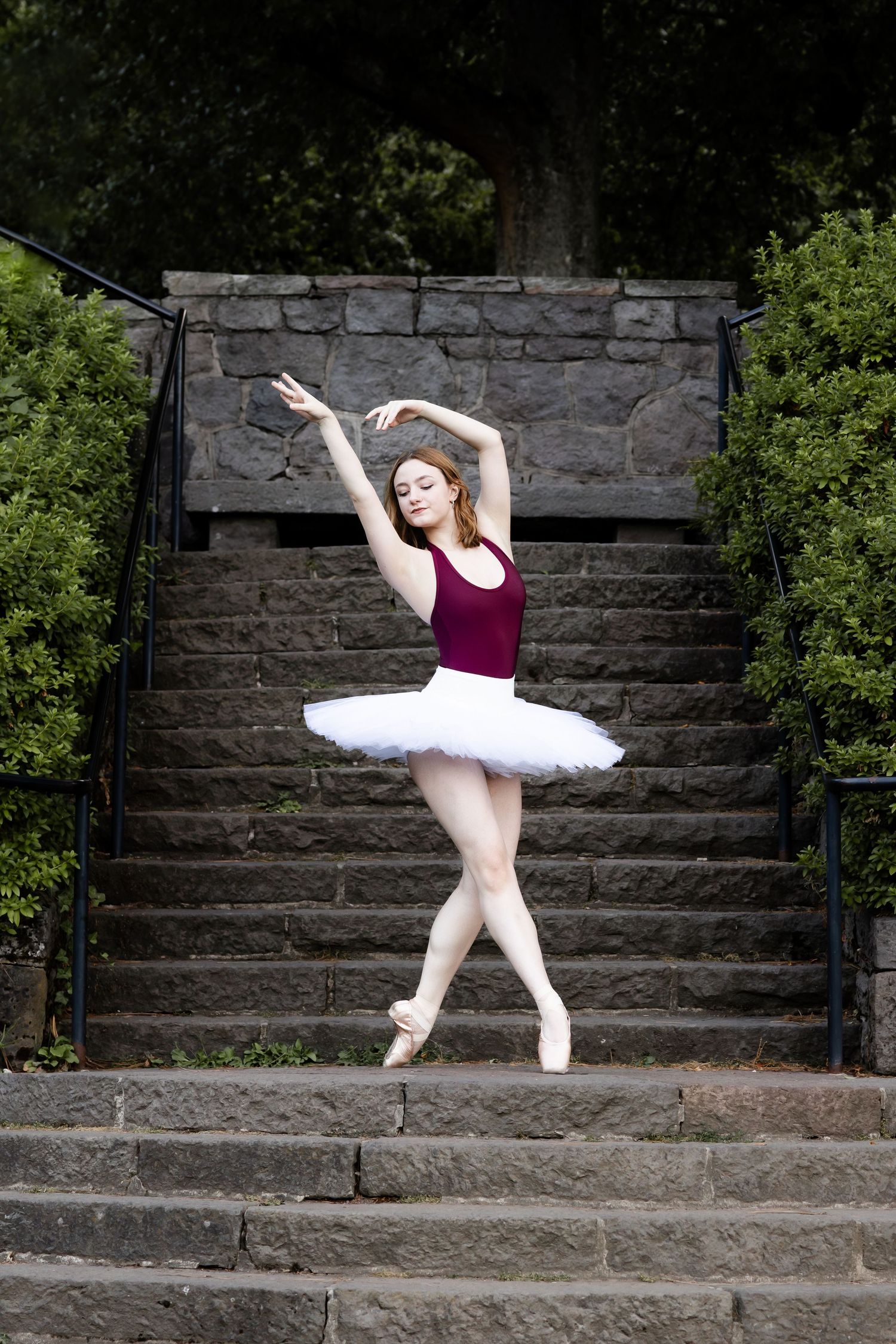 A ballet dancer in a white tutu and burgundy leotard performs an elegant pose on stone steps surrounded by foliage.