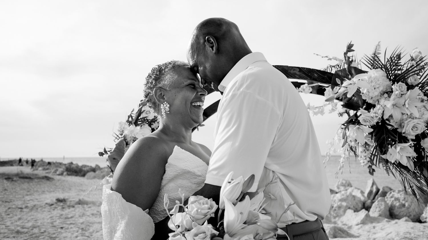 Black and white beach wedding photo of couple sharing romantic moment near floral arrangement.