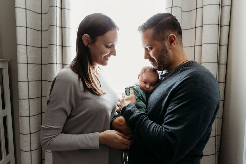 New parents lovingly cradling their newborn baby together by a bright window in a tender family portrait.