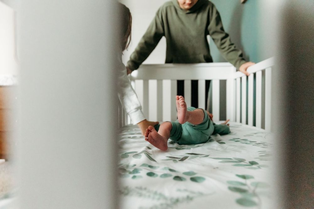 Parent leaning over white crib watching newborn baby lying on floral mattress in green outfit.