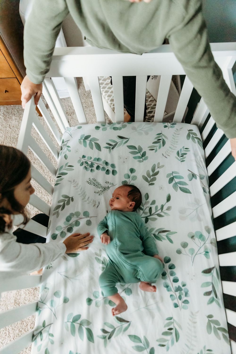 Two parents lovingly look down at their newborn baby lying in a white crib with a green leaf patterned sheet.