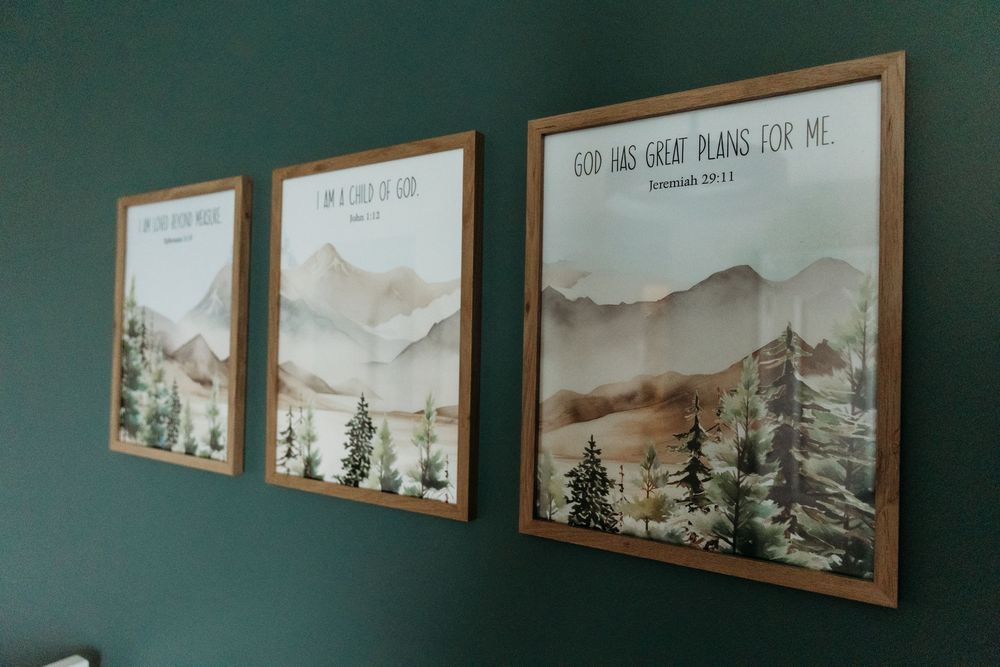Three framed mountain landscape prints with inspirational Bible verses hang on a dark green wall with wooden frames.