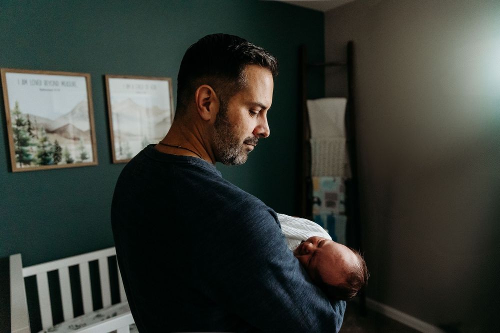 A father tenderly holds his newborn baby in a softly lit nursery room with green walls.