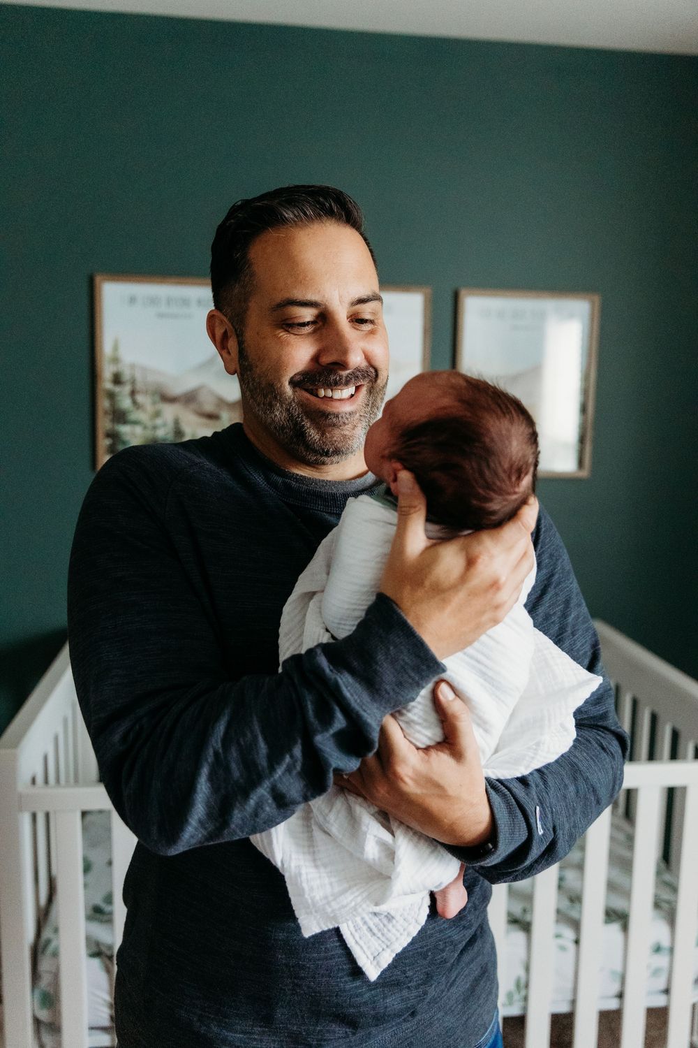 Smiling father holding newborn baby in nursery with green walls and white crib.