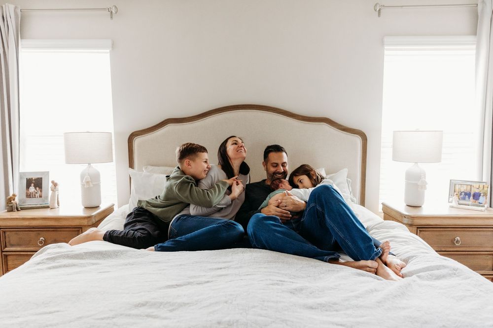 Happy family of four cuddling together on a white bed in a bright, cozy bedroom with natural light.