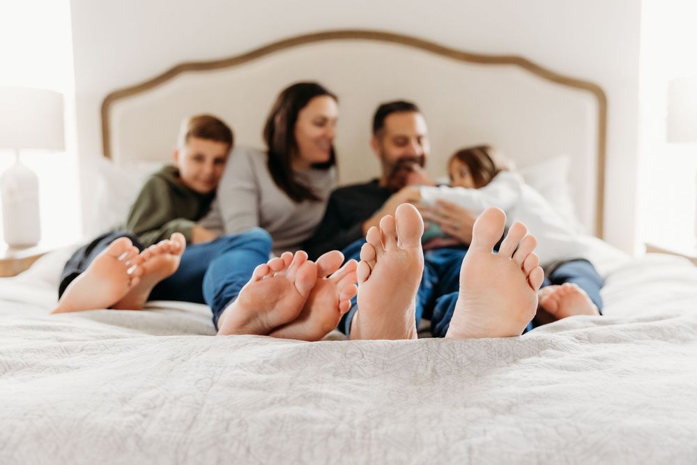 Family of four relaxing on a white bed with bare feet in foreground, parents and children enjoying a cozy morning together.