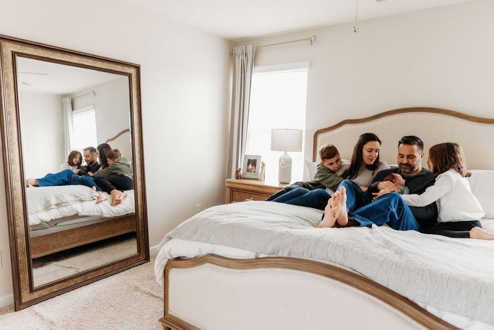 Family of four relaxing together on a white bed in a bright bedroom with a large floor mirror reflecting their cozy moment.