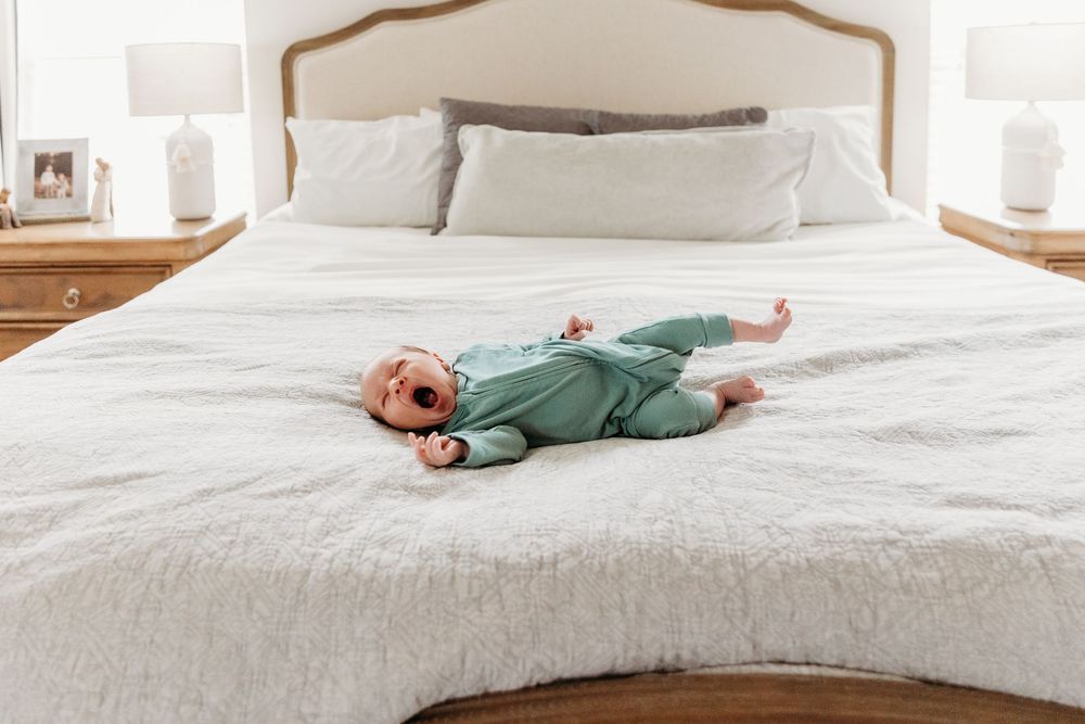 A baby in a green onesie lying on a large white bed with neutral bedding and wooden nightstands.