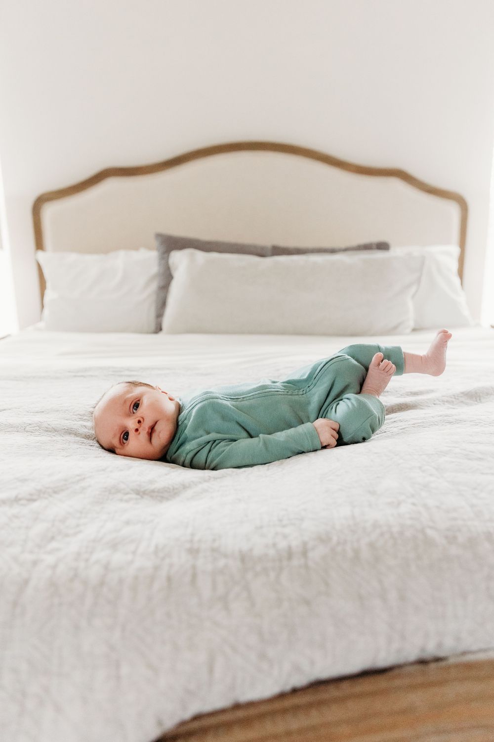 Cute baby in green onesie lying on a white bed with plush bedding and upholstered headboard.
