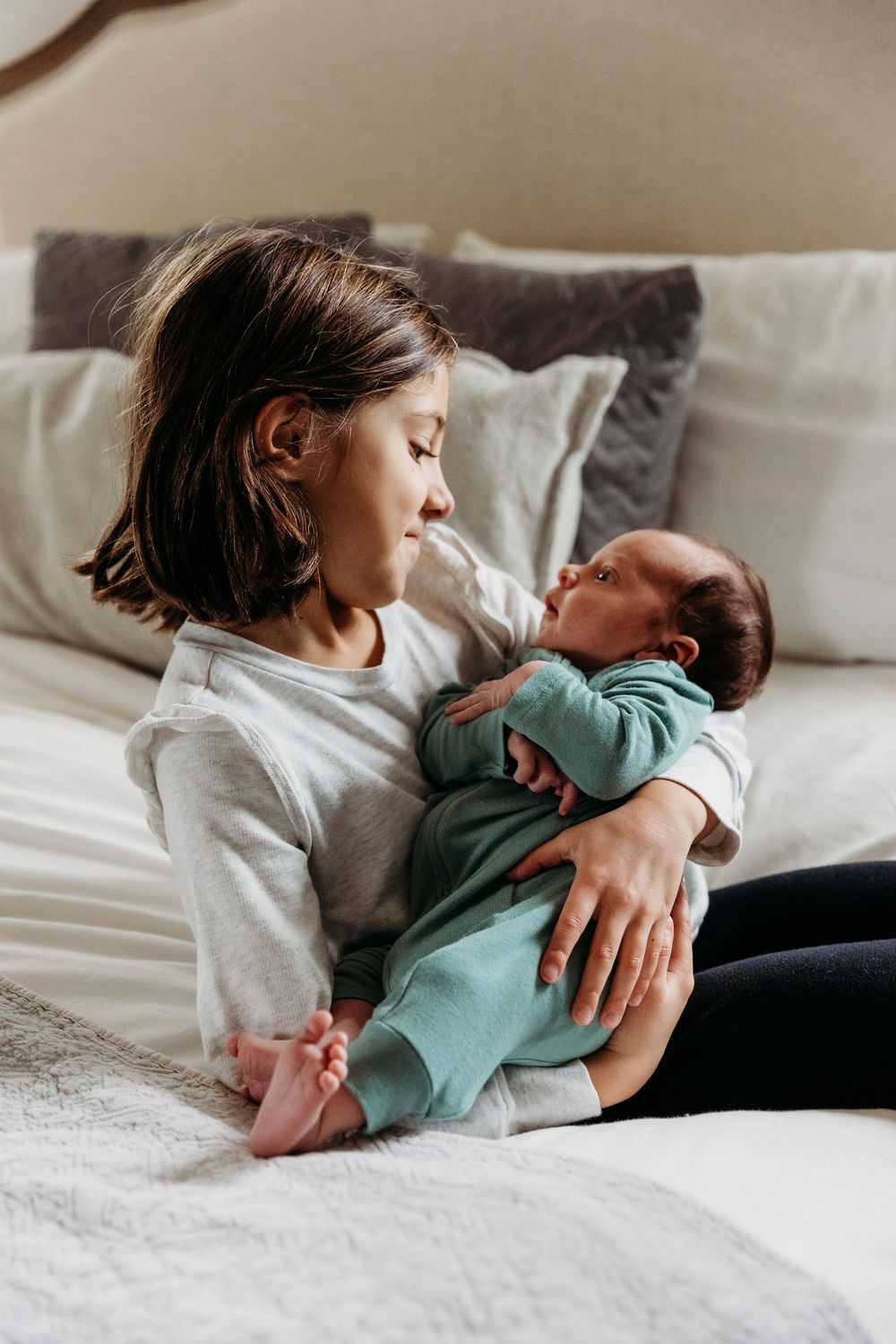 A young girl sits on a bed cradling a newborn baby dressed in a teal outfit against soft white pillows.