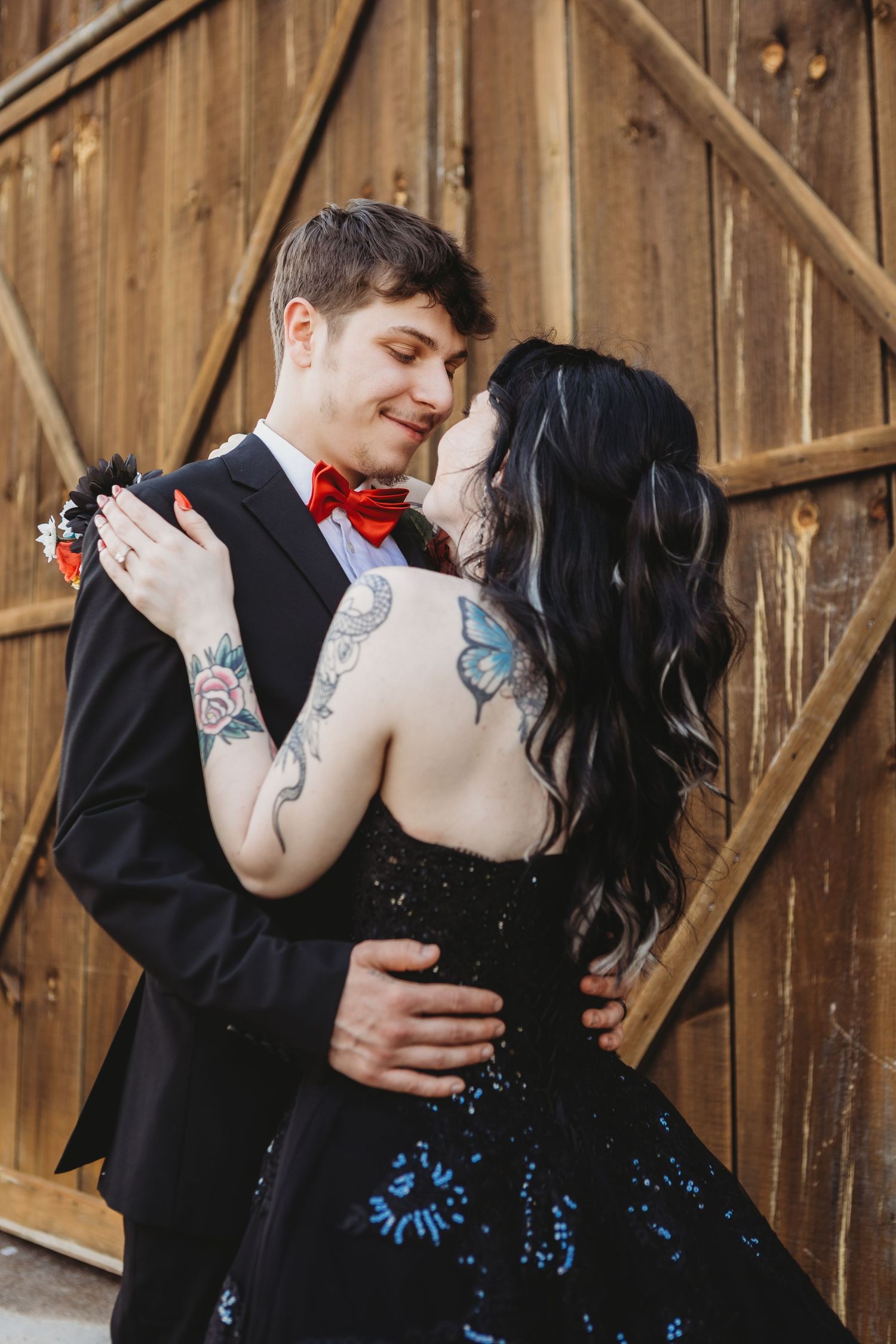 A tattooed couple in formal attire embraces romantically in front of a rustic wooden barn door.