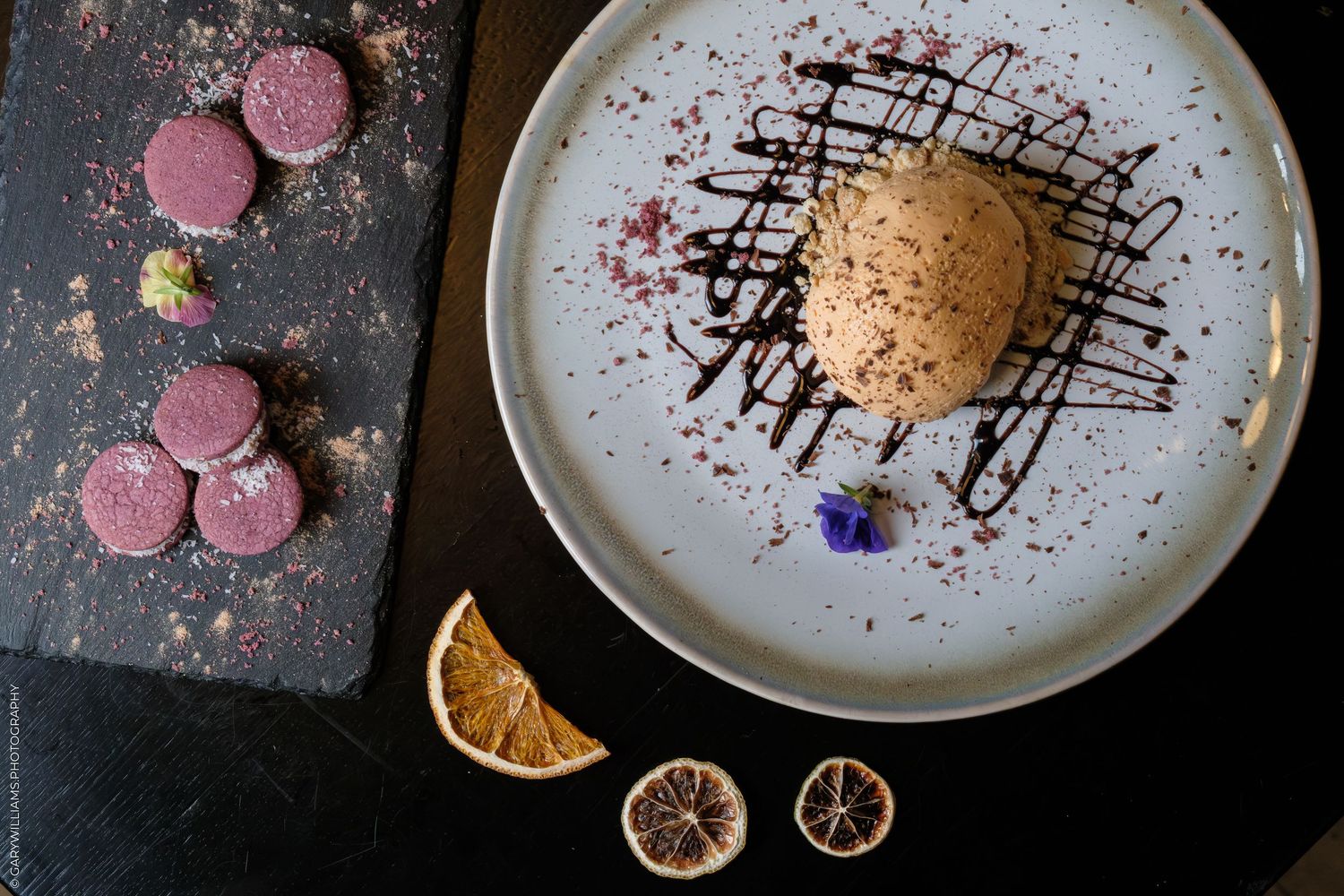 Elegant dessert plate with chocolate ice cream, drizzle, edible flower, macarons on slate, and dried orange slices.
