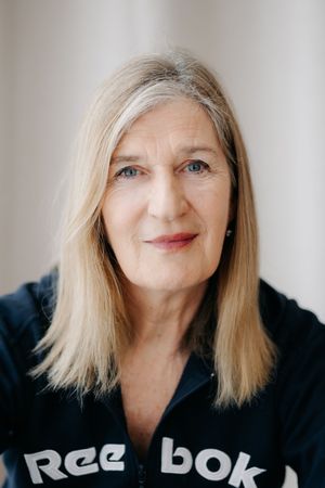 Older woman with blonde hair wearing a Reebok jacket, portrait shot against a light background.