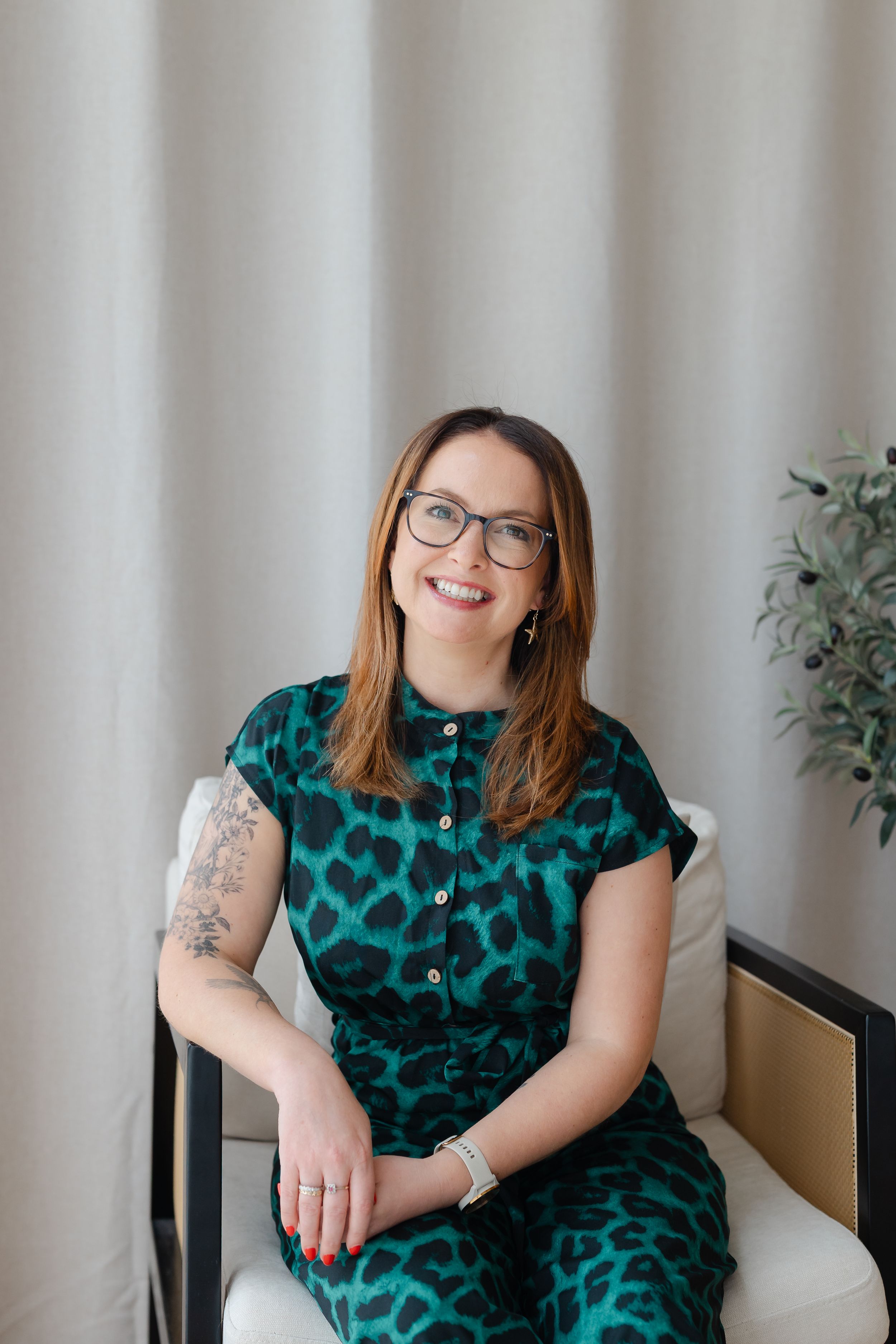 Smiling woman with glasses wearing a green leopard print dress, seated in a chair against neutral curtains.