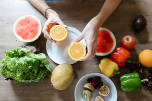 Hands holding a halved orange over a white plate surrounded by fresh fruits and vegetables on a wooden table.