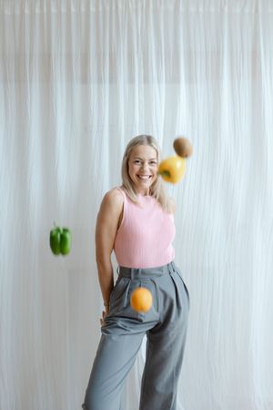 Smiling blonde woman in pink top and gray trousers posing with flying fruits and vegetables against white curtain.