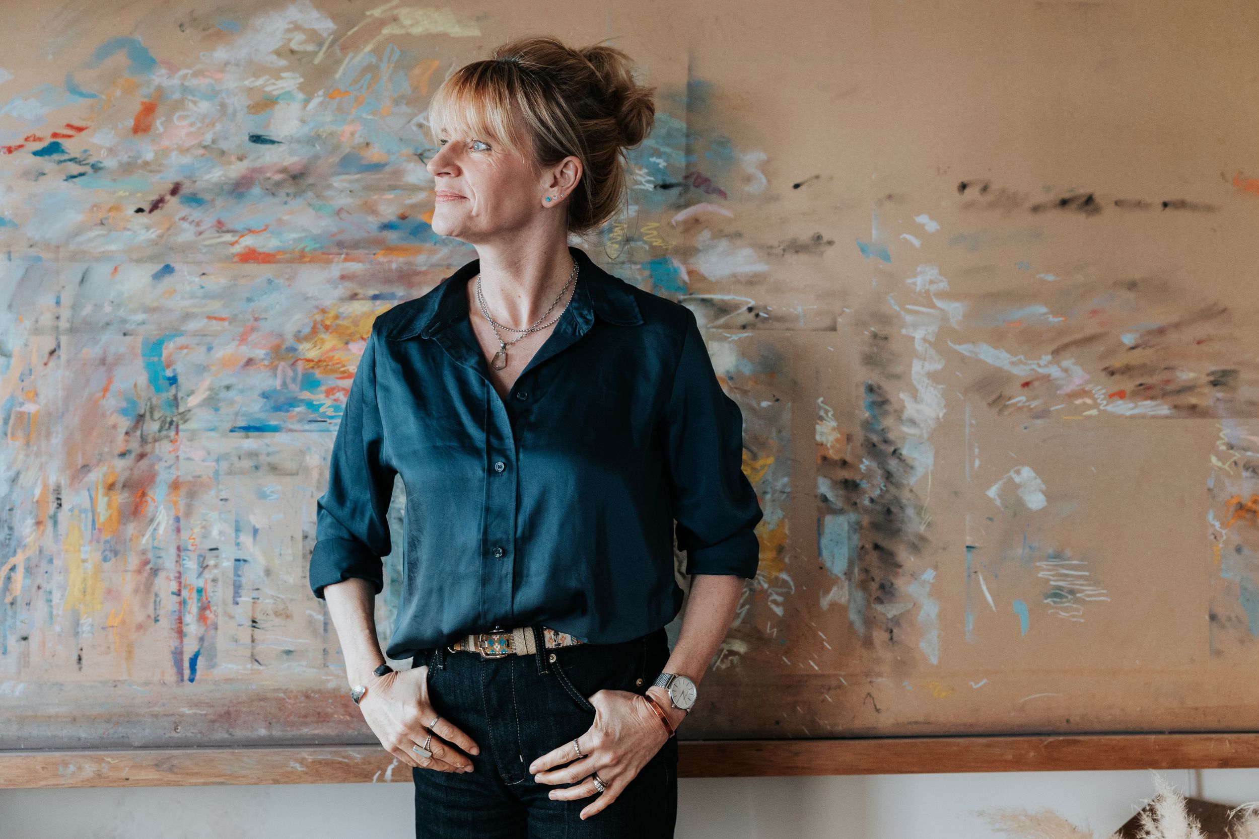 Someone in a navy blue button down shirt and dark jeans poses casually against a weathered wall with peeling paint.