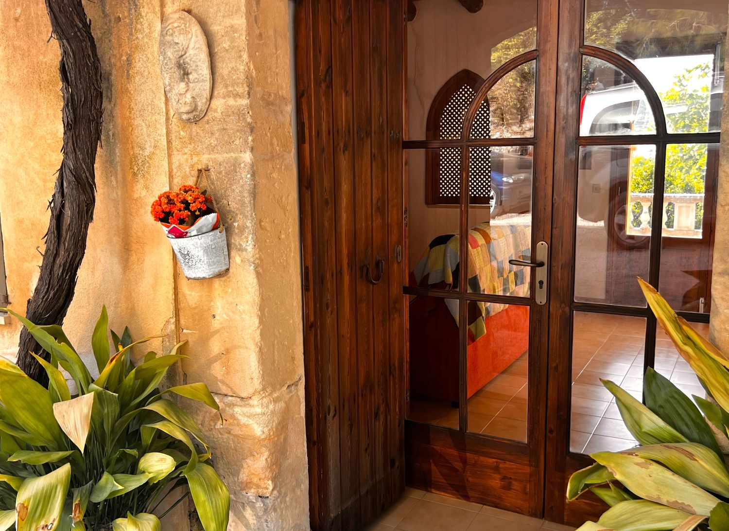 Rustic stone doorway with wooden arched door, potted orange flowers, and tropical plants at a Mediterranean villa entrance.