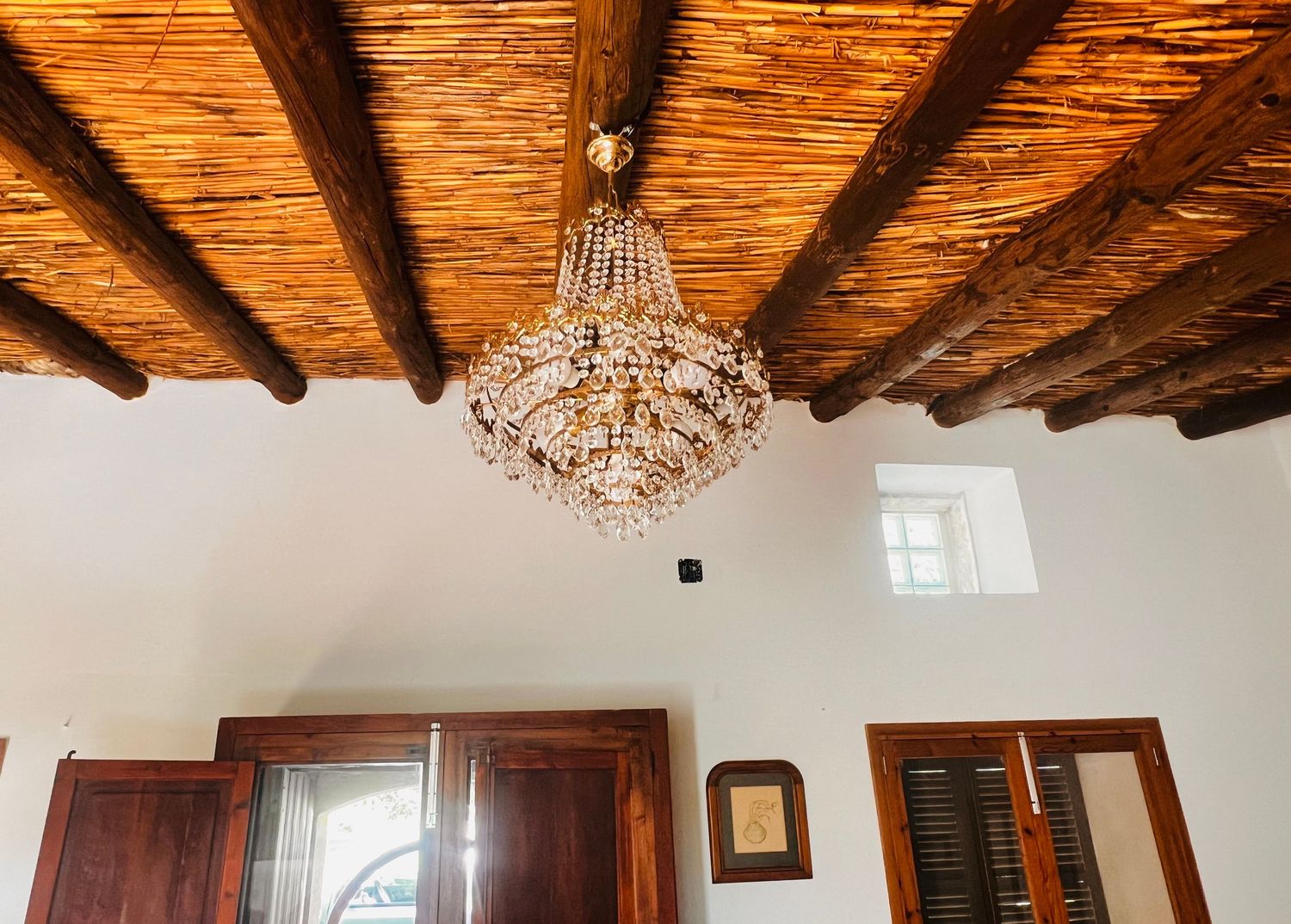 Elegant crystal chandelier hanging from rustic wooden beam ceiling with mahogany doors in a Mediterranean style room.