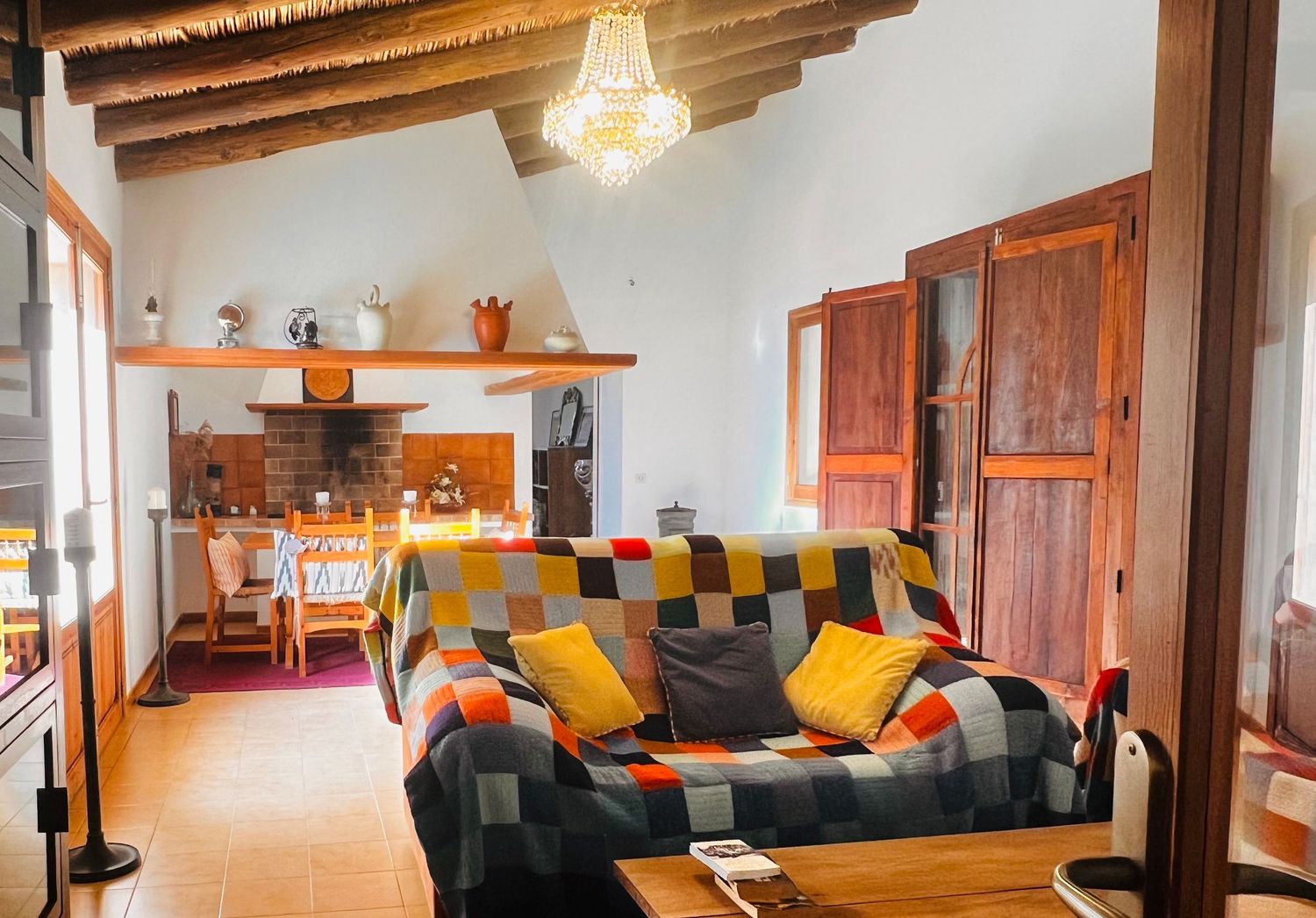 Cozy rustic living room with colorful patchwork sofa, wooden beamed ceiling, chandelier, and open kitchen dining area.