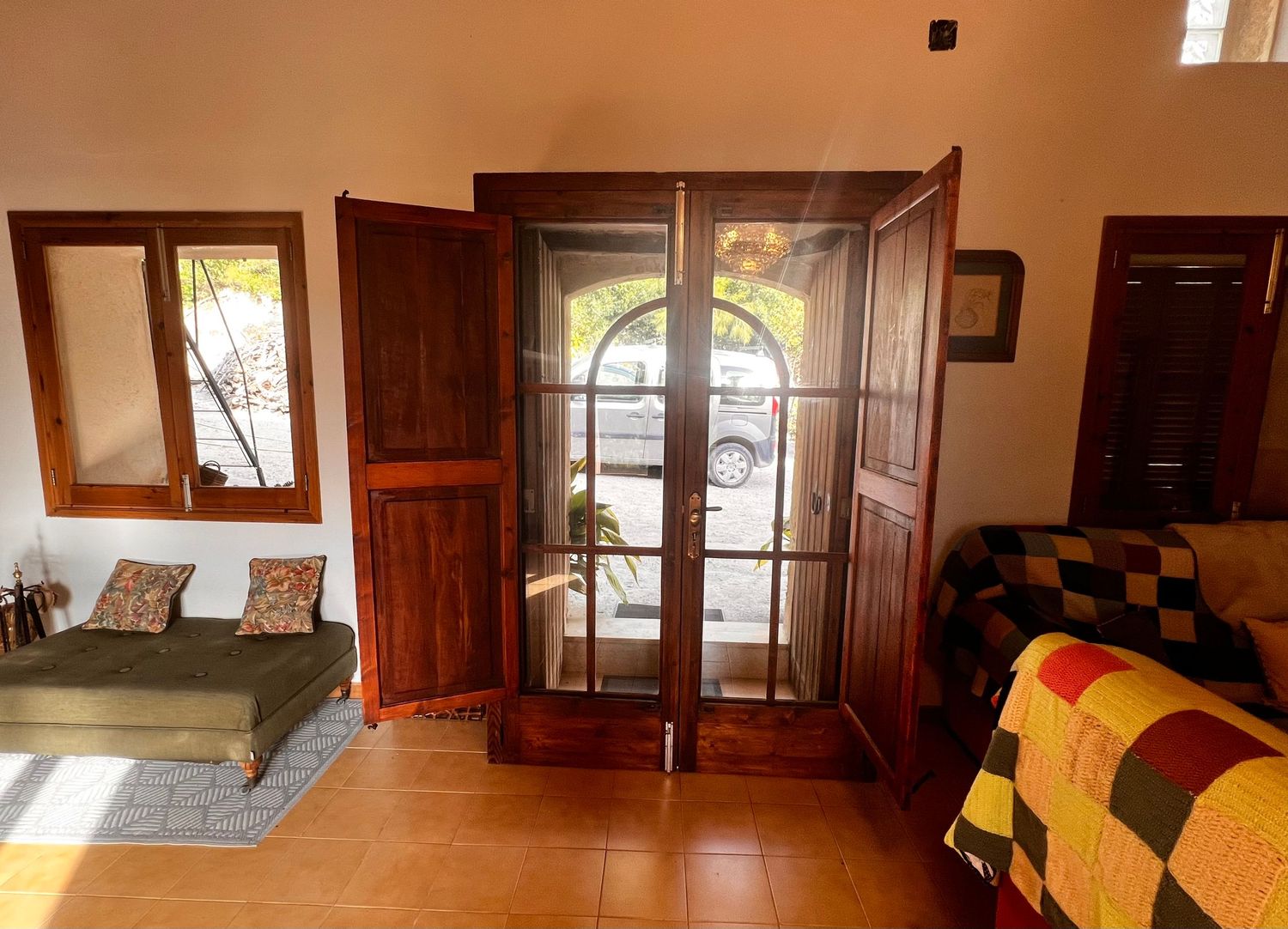 Sunlit room with open wooden French doors leading to a patio, tiled floors, a sofa, and a colorful patchwork bedspread.