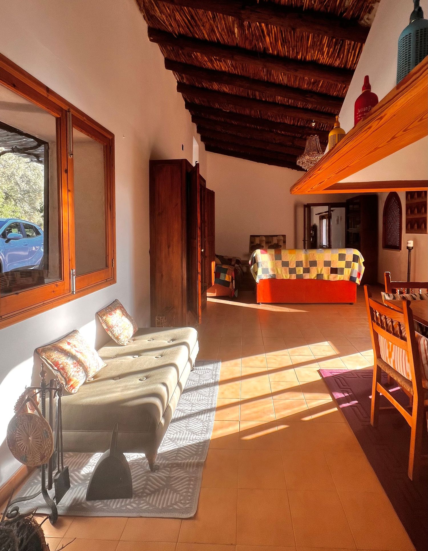 Sunlit rustic interior with terracotta floors, wooden beam ceiling, lounge chairs, and colorful bedding in a Mediterranean-style villa.