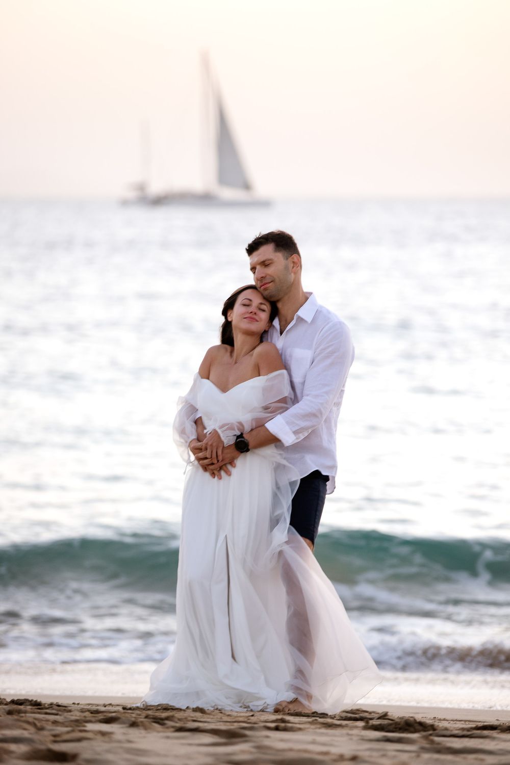 Romantic couple embracing on a sandy beach at sunset, woman in white dress, sailboat in background.