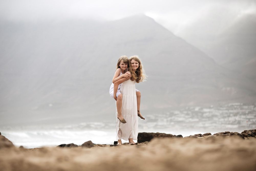 Woman in white dress giving a child a piggyback ride on a rocky beach with misty mountains in the background.