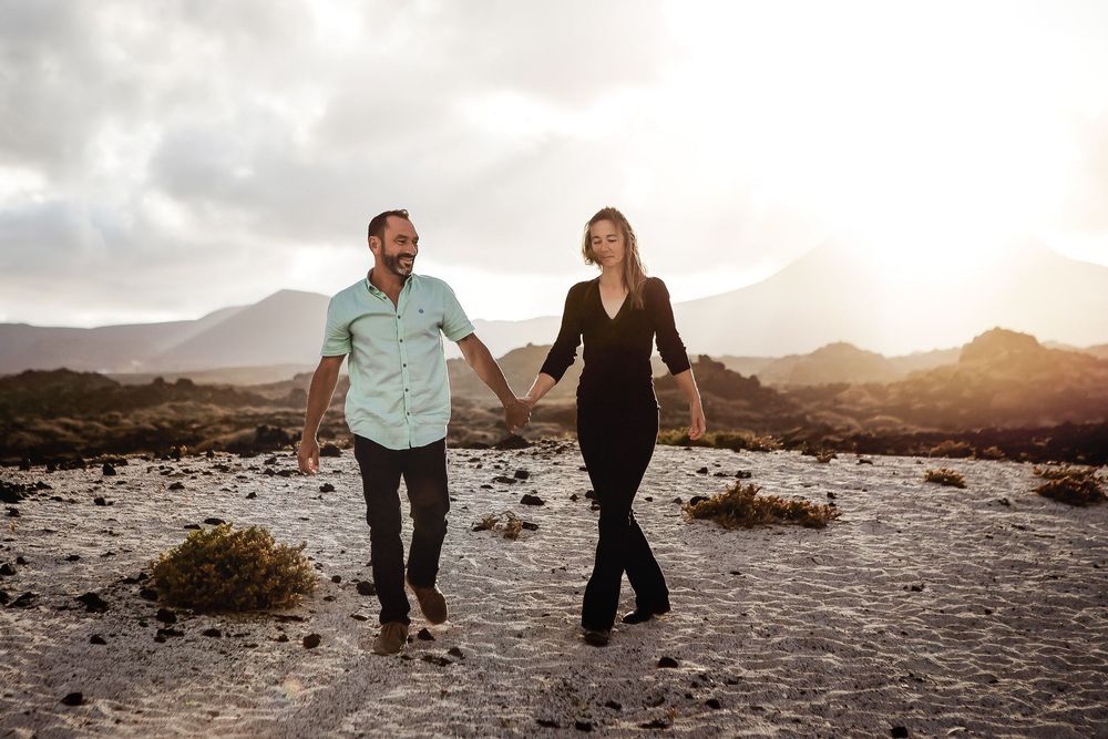 Couple holding hands walking through volcanic desert landscape with mountains and golden sunset in background.