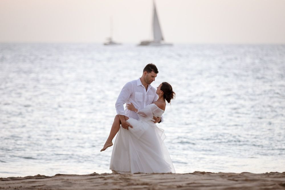 Romantic couple in white attire sharing a tender embrace on a sandy beach with a sailboat in the background.