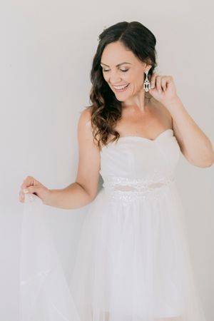 Happy brunette bride in strapless white wedding dress smiling while putting on chandelier earrings before ceremony.
