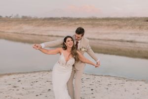 Joyful bride and groom dancing playfully on sandy beach at sunset near a calm river.