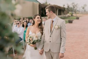 Smiling newlyweds walk hand-in-hand past guests after outdoor wedding ceremony on brick pathway.