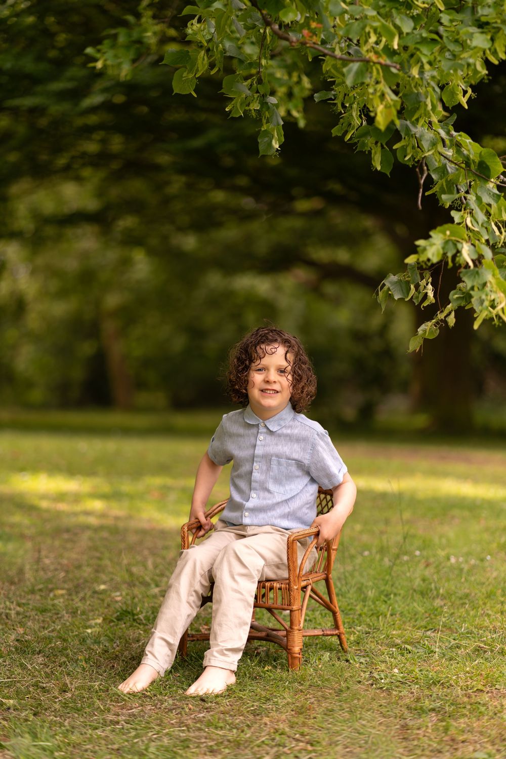 A curly-haired boy sits smiling on a wicker chair outdoors on green grass beneath leafy trees.
