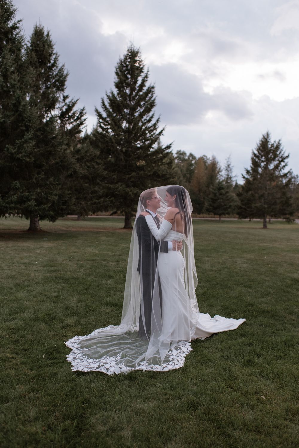 Bride and groom sharing a kiss under a long lace-trimmed veil on a green lawn surrounded by evergreen trees.