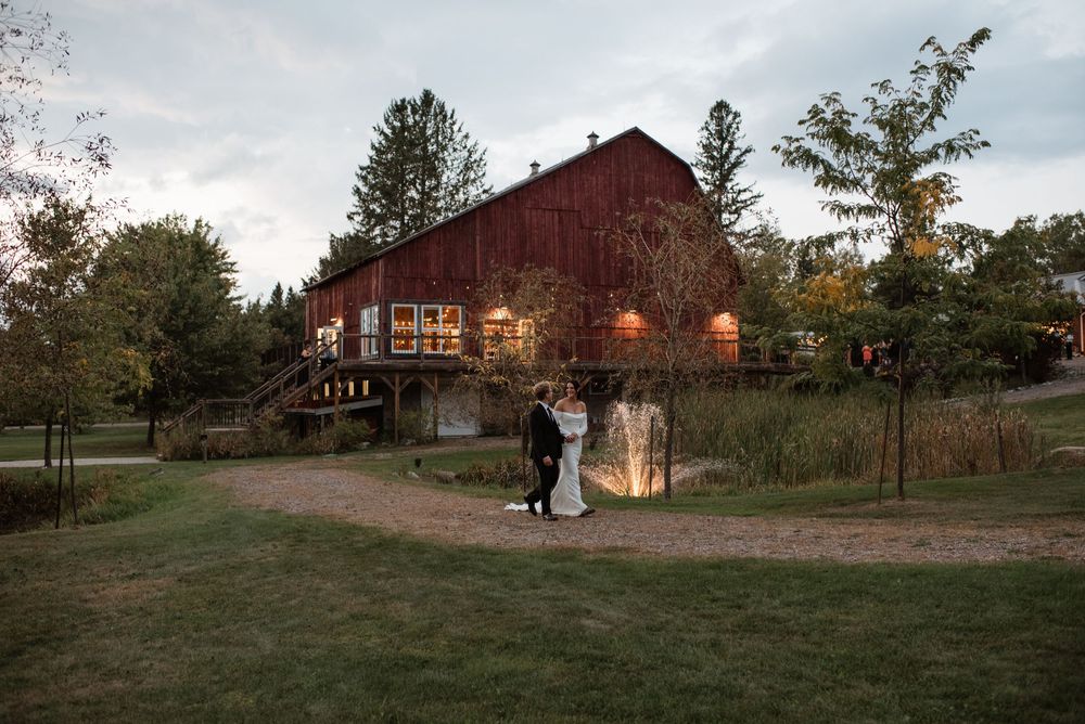 Bride and groom embrace near fountain outside illuminated red barn wedding venue at twilight.
