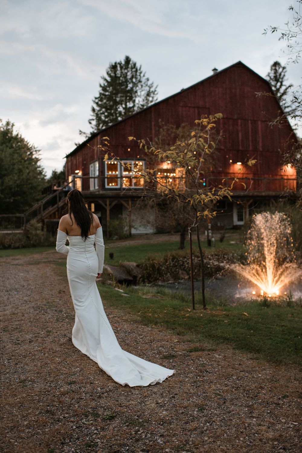 Bride in white gown with train walks toward lit red barn venue at dusk with fountain nearby.