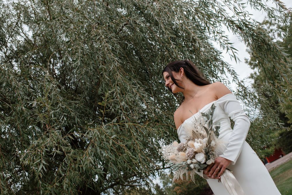 Bride in elegant off-shoulder white gown smiling while holding a boho bouquet beneath a willow tree.