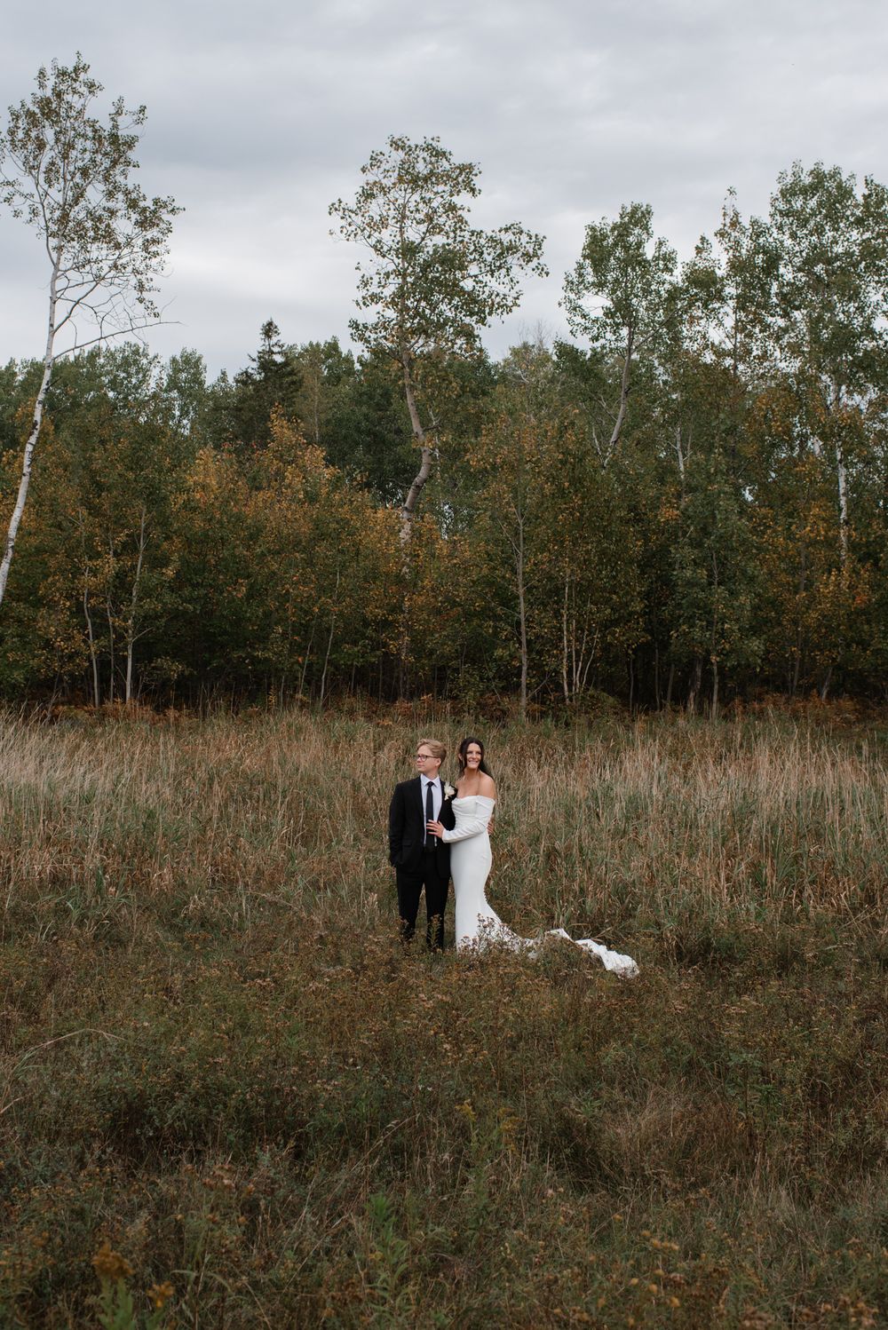 Bride and groom standing in an autumn field with golden trees and overcast skies on their wedding day.