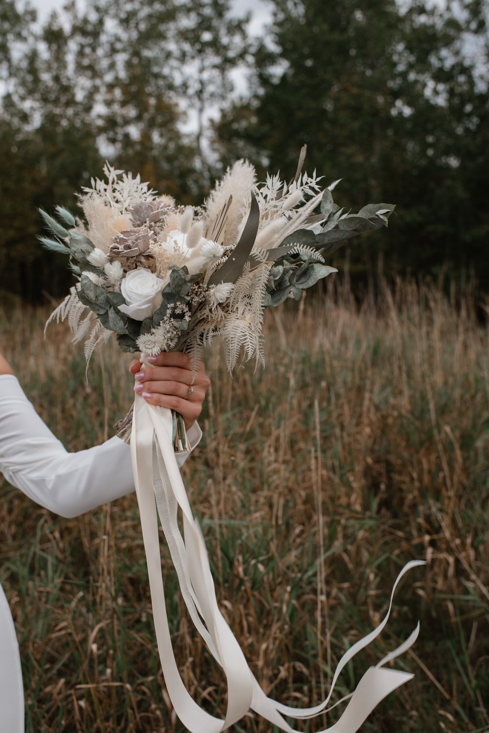 Bride holds a boho wedding bouquet with white roses, pampas grass, and ribbon streamers in a field.