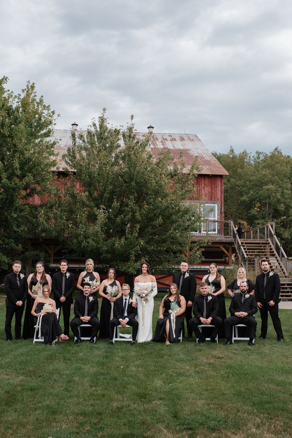 Wedding party in black attire poses on green lawn before a rustic red barn on a cloudy day.