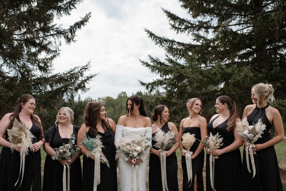 Bride in white gown laughs with bridesmaids in black dresses holding boho bouquets under pine trees.