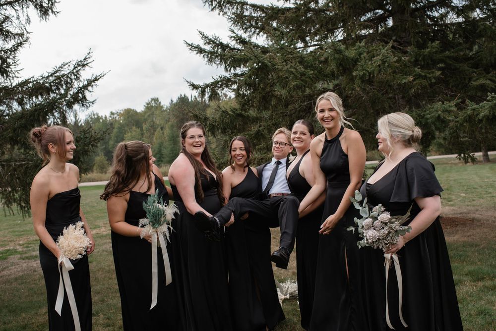 Bridesmaids in black gowns playfully lift groom during fun outdoor wedding party photo session.
