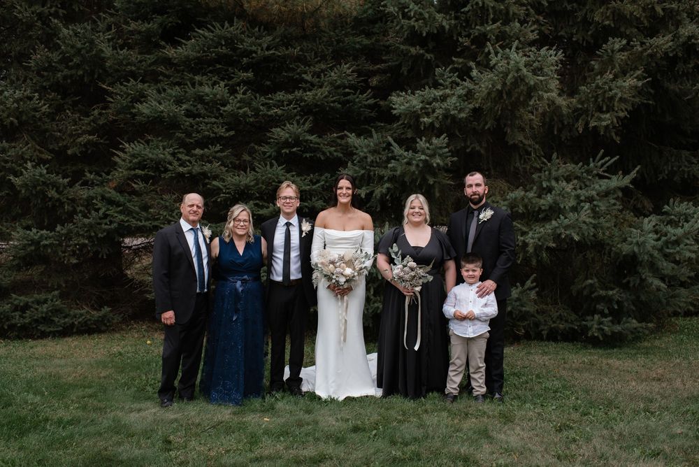 Wedding party group portrait with bride and groom surrounded by family members in front of evergreen trees.