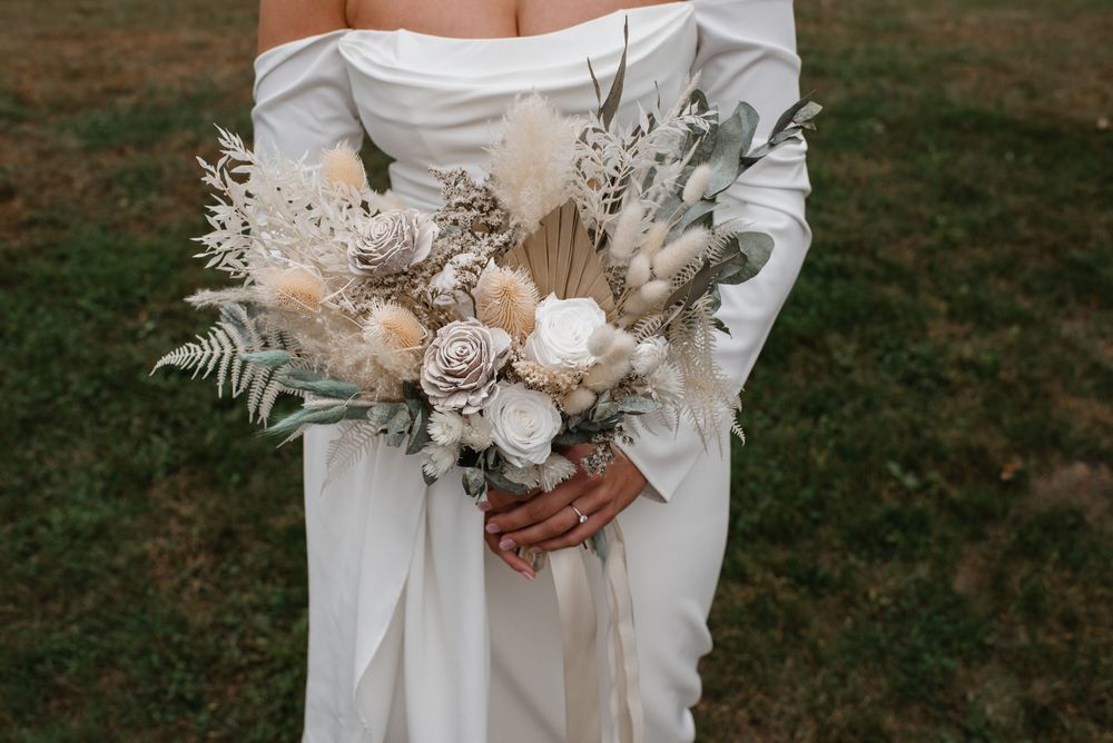 Bride holding a boho wedding bouquet with dried pampas grass, white roses, and neutral toned flowers in a white gown.
