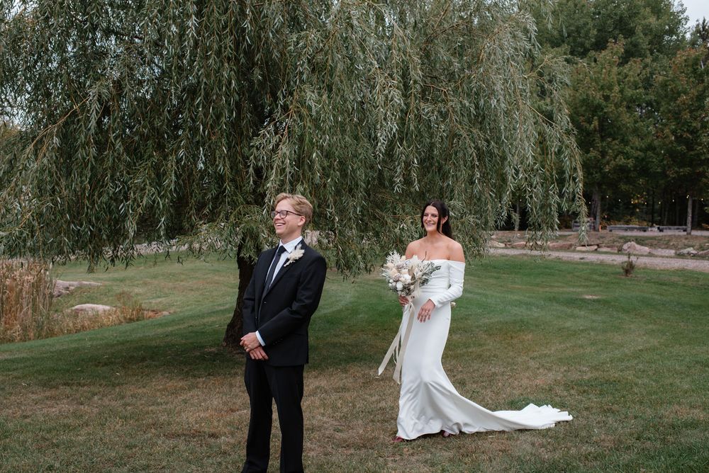Bride and groom share a first look moment on a lush green lawn beside a weeping willow.