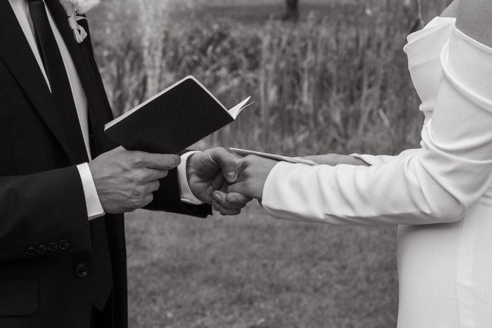 Black and white close-up of groom holding vow book while bride and groom hold hands at ceremony.