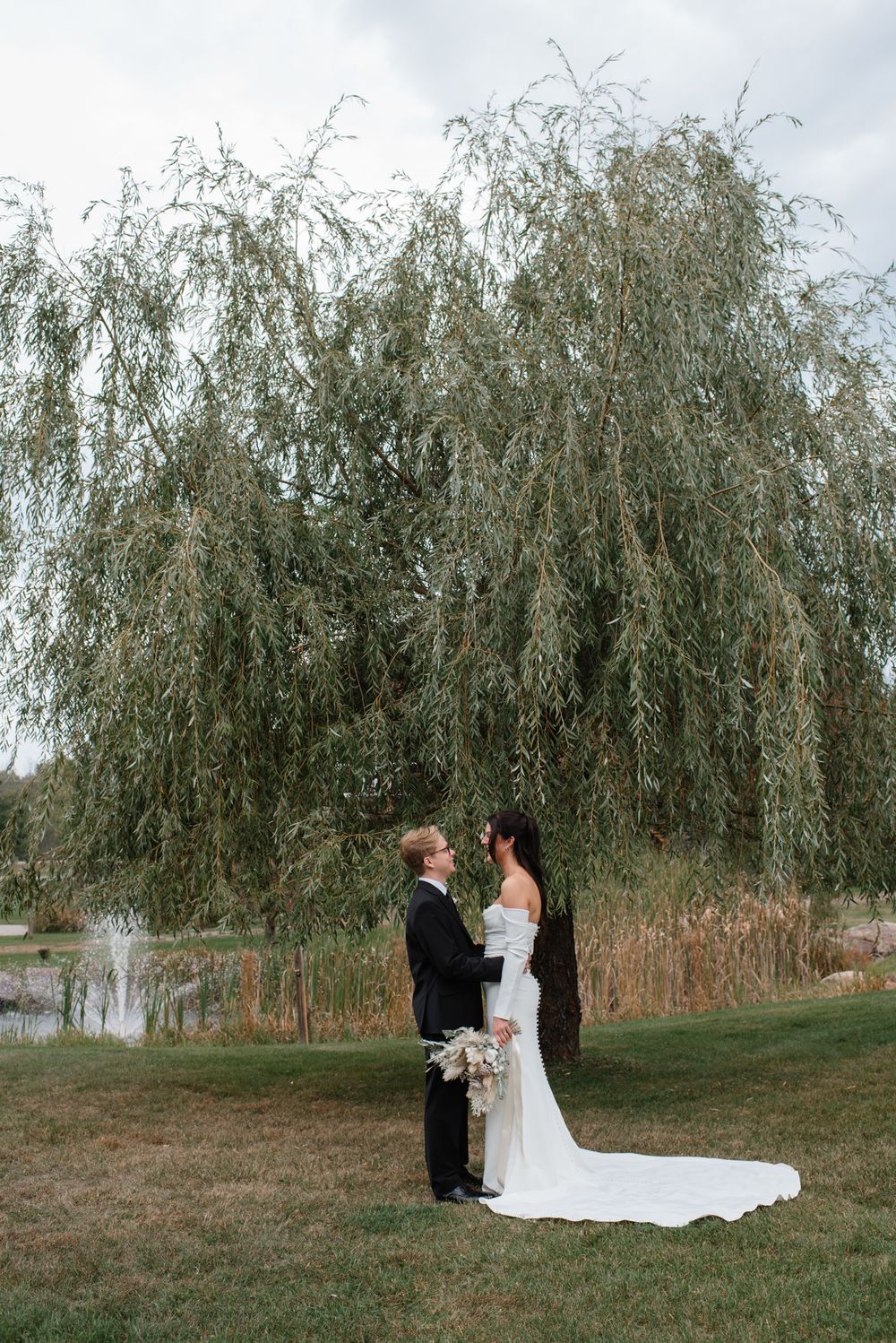 Bride and groom face each other holding hands under a weeping willow tree beside a pond.